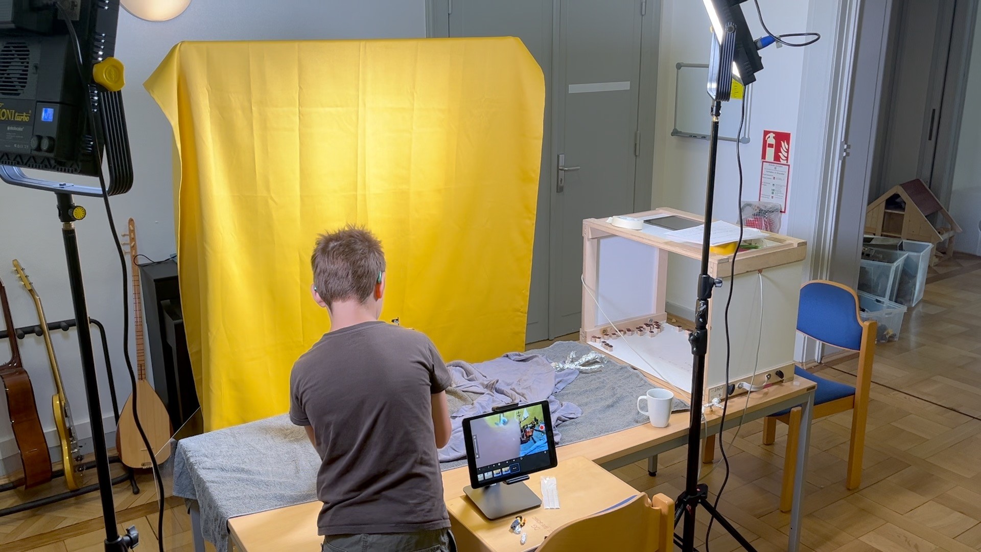 Ein kurzhaariges Kind steht mit dem Rücken vor einem Tisch darauf steht ein Tablet hinter dem Tisch ist ein glebes Tuch an der Wand gespannt. Links und rechts vom Tisch sind Fotoleuchten auf Stativen.