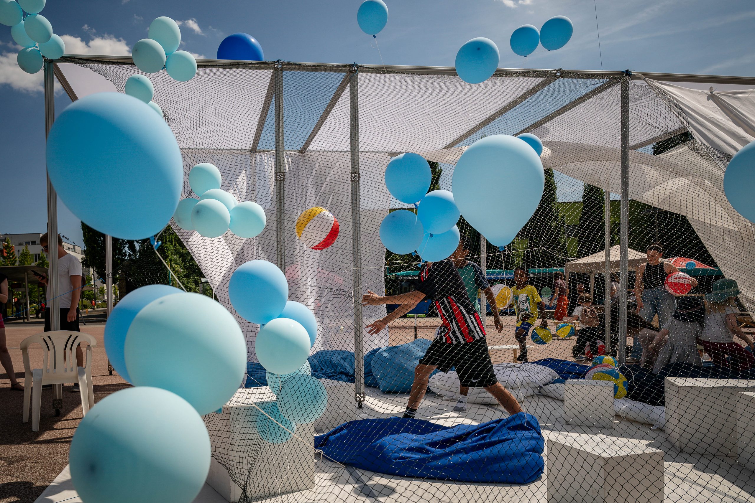 Hellblaue Luftballongs fliegen vor einem Netz. Darin Springt ein Kind. Der Himmel ist auch hellblau.