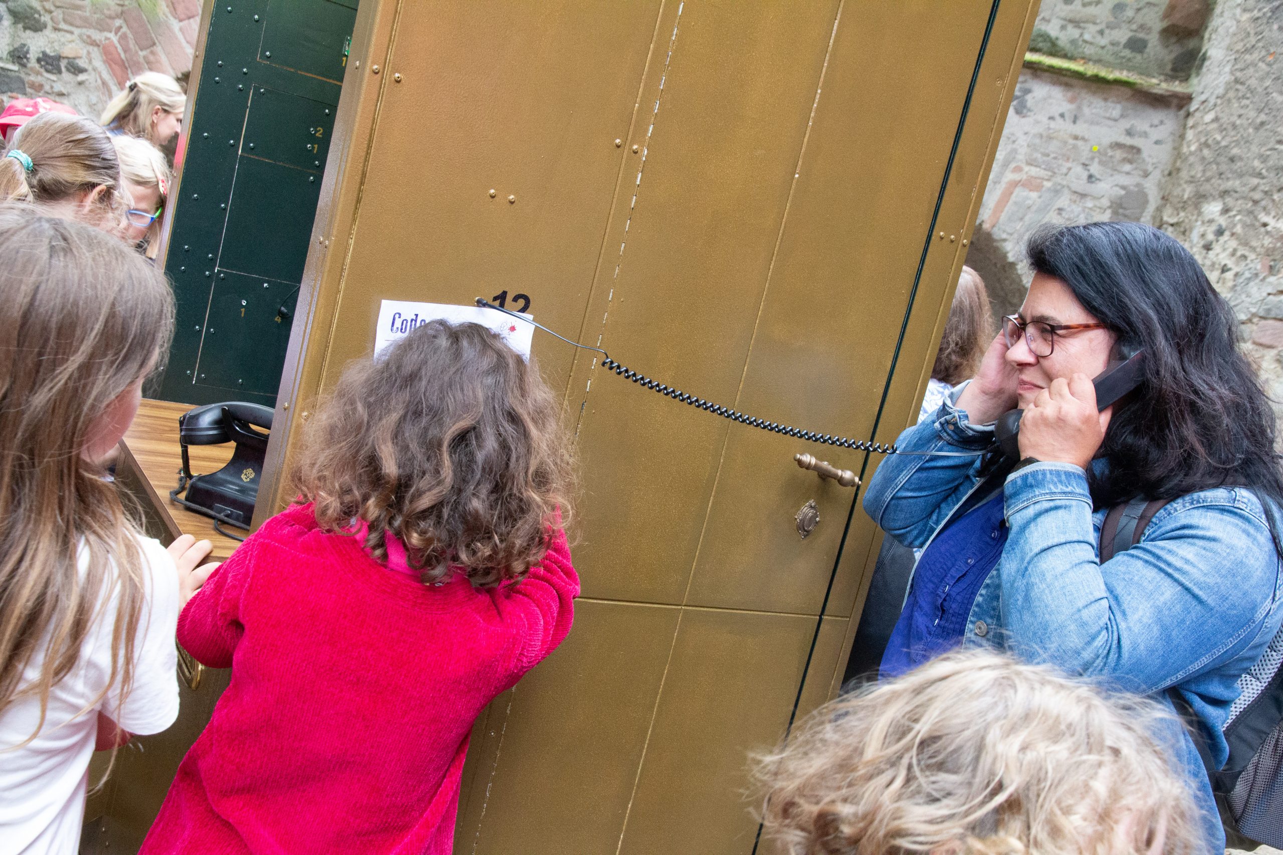 Links zwei Kinder von hinte. Rechts im Profil eine Frau mit braunen Haaren, die ein Telefonhärer mit Schnur hält.