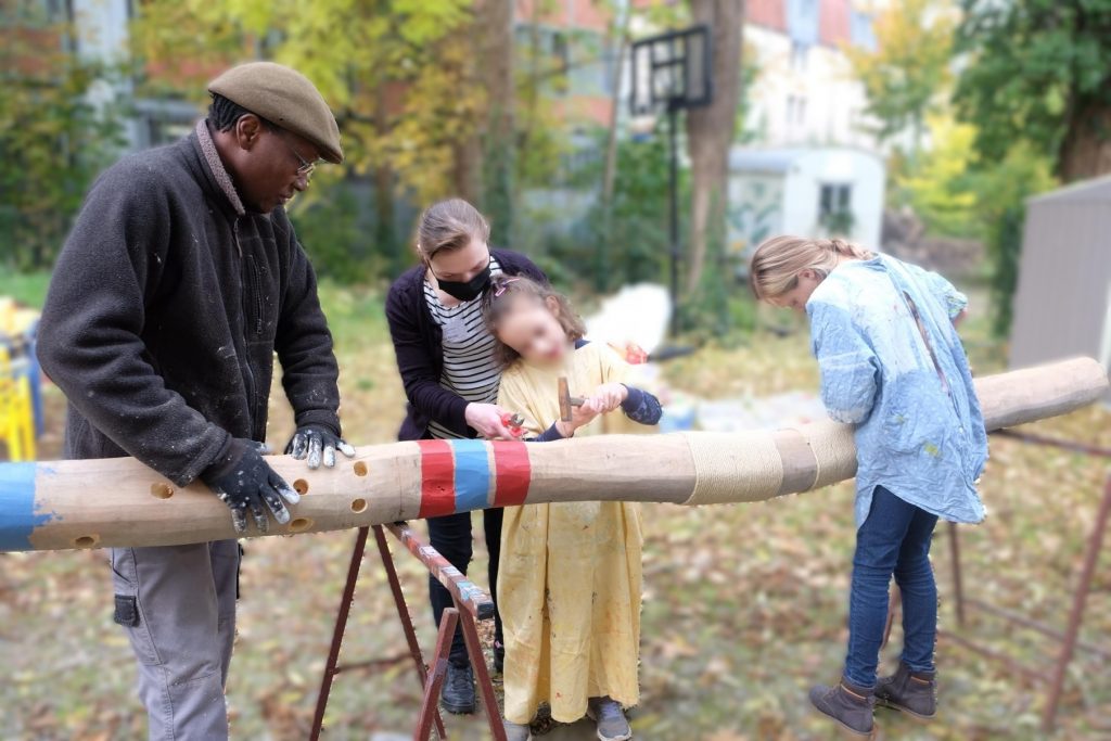 Ein Kind steht hinter einem Baumstamm waagerecht aufgebockt. links und rechts stehen Erwachsene.