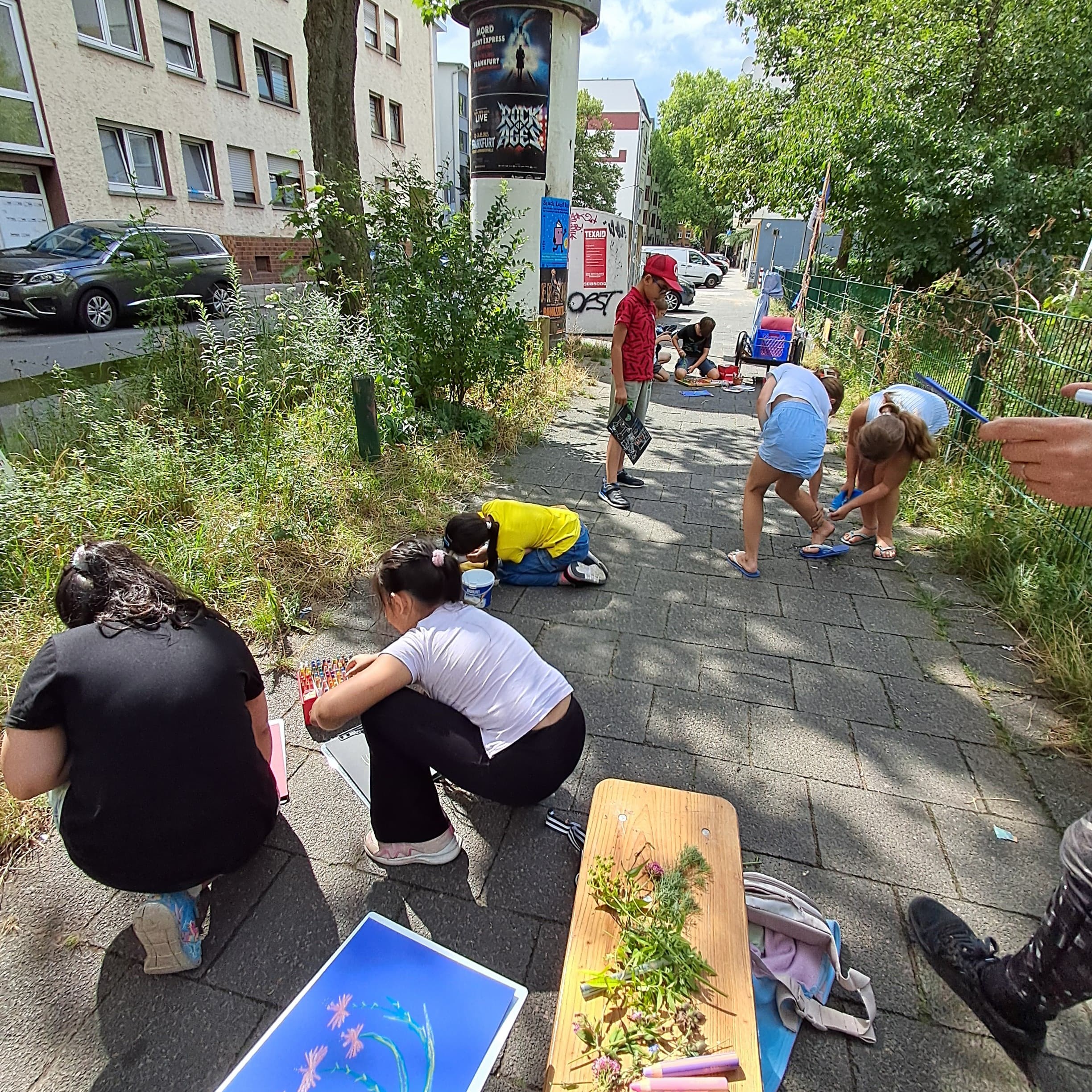 Auf einem Gehweg mit rechts und links Grünstreifen, hocken Kinder mit dem Rücke zu der Kamera. Auf einer Bank liegen Farben.