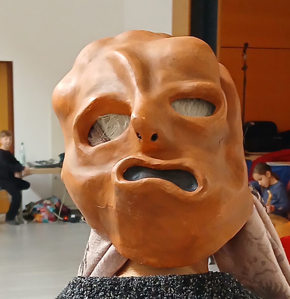 Eine beigbraune Gesichtsmaske. Dahinter ein Raum mit weißerwand und Holztür.