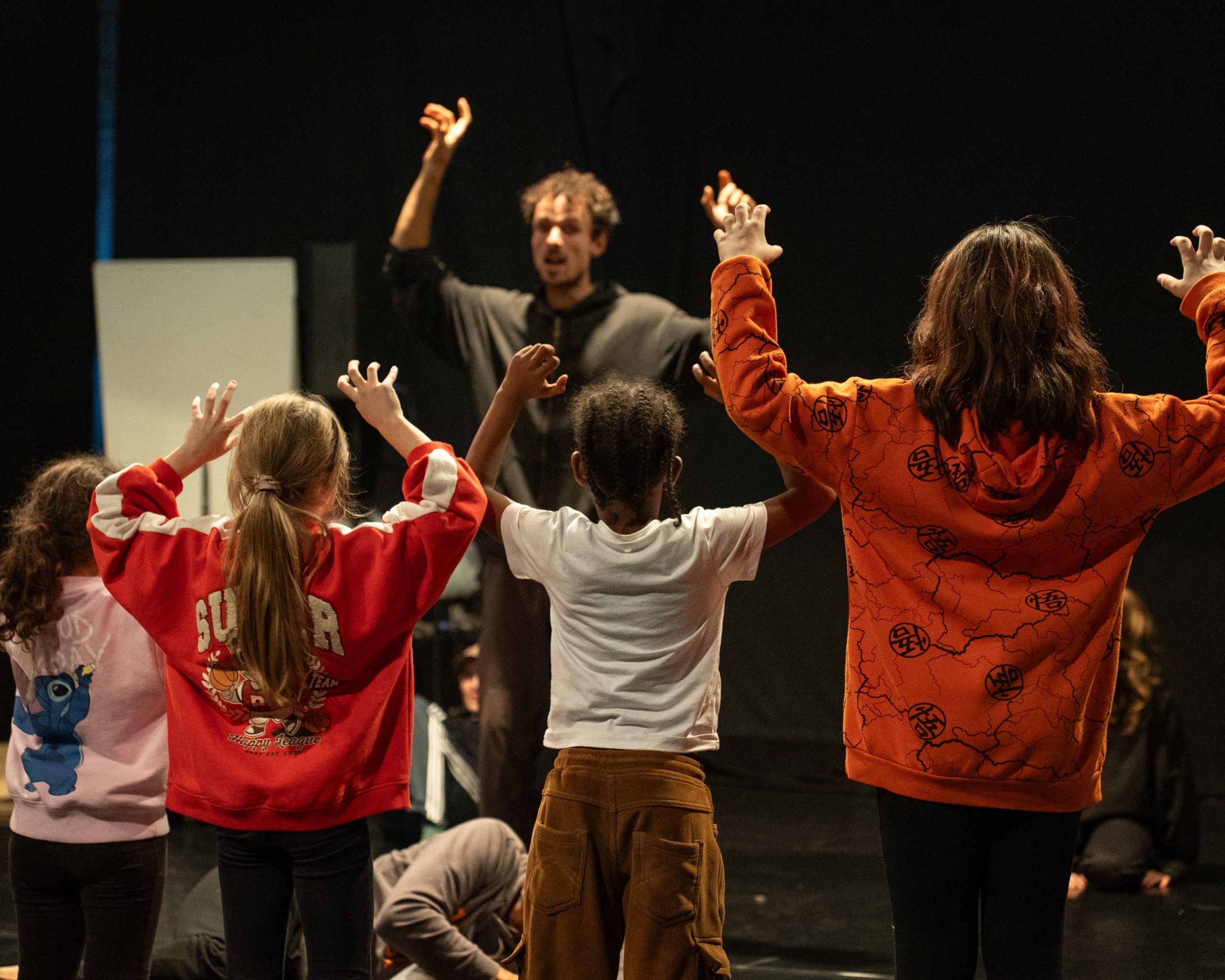 Drei Kinder von Hinten heben die Hände alaog zu einem Mensch mit gehobenen Händen der Richtung der Kamera schaut.