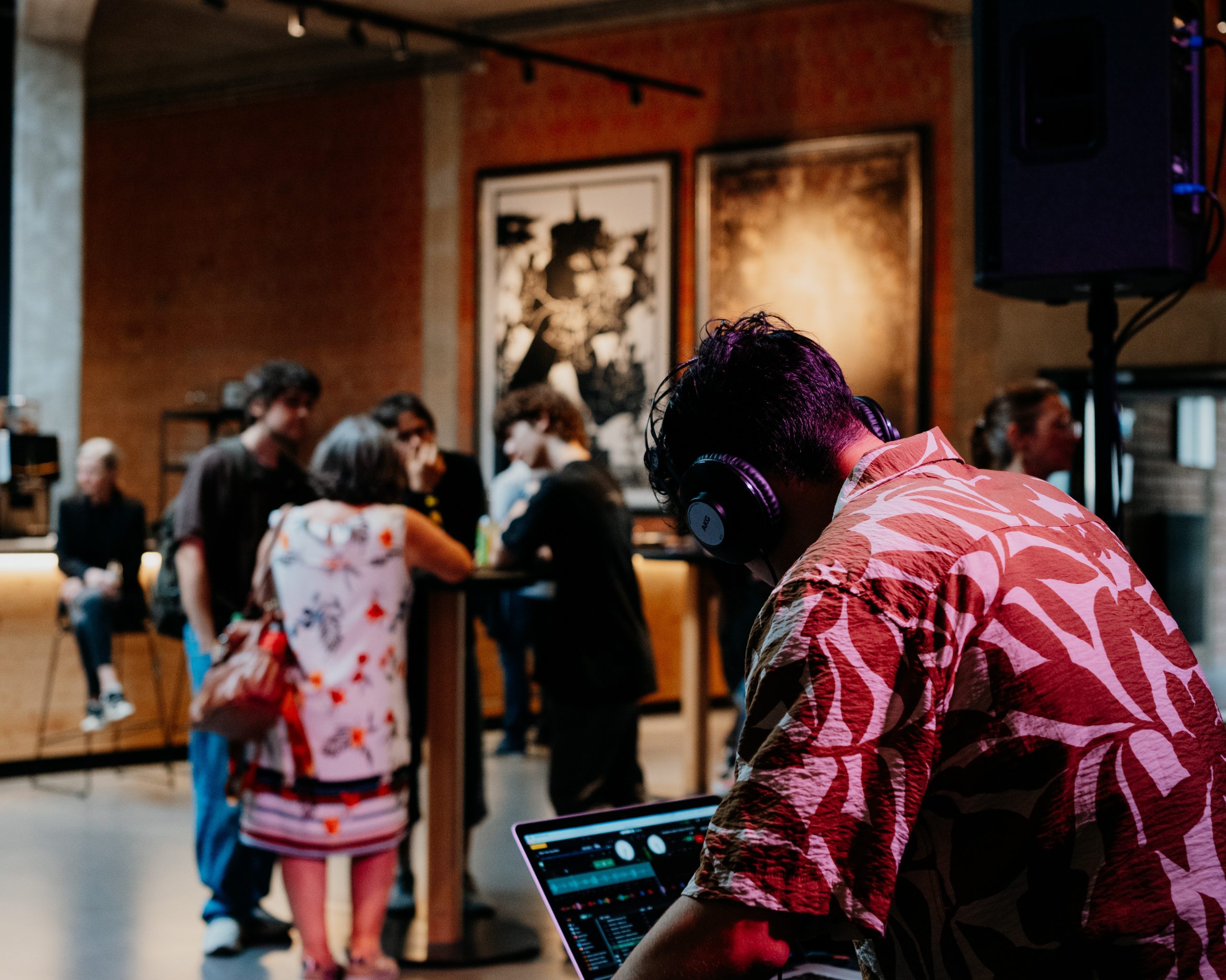 Im Vordergrund links steht seitlich von hinten ein Mensch mit schwarzen Haaren und Kopfhörer vor einem Laptop. Im Hintergrund stehen eine Gruppe von Menschen zusammen.