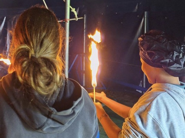 Zwei Kinder mit dem Rücken zur Kamera. Ein Kind hält einen Holzstab mit einer Feuerflamme.