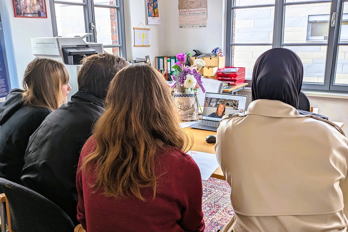 Vier Menschen sitzen mit dem Rücken zur Kamera vor einem Tisch auf dem ein Latop liegt. Auf dem Bildschirm des Laptops ist eine Bildschirmkachel mit einer Frau zu sehen.