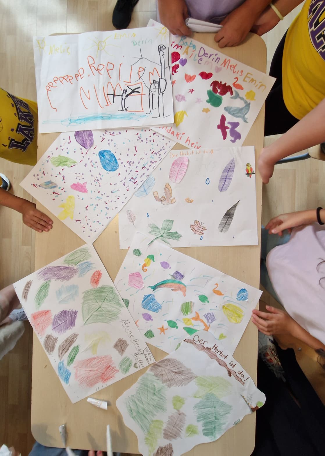 Ein Tisch auf dem viel selbstbemalte Papiere zu sehen sind wird aus der Vogelperspektive eingefangen. Kinderarme sind am Rand sichtbar. Sie sitzen um den Tisch herum.